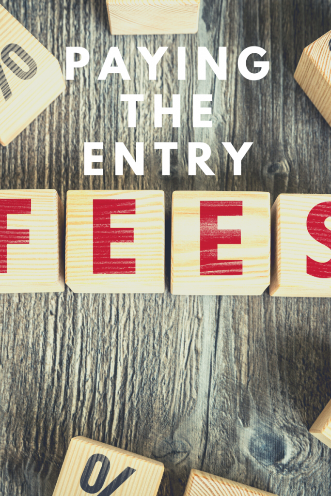 On a wooden table are wooden blocks with letters on them spelling out fees. The text reads paying the entry above the word fees so it reads paying the entry fees. 