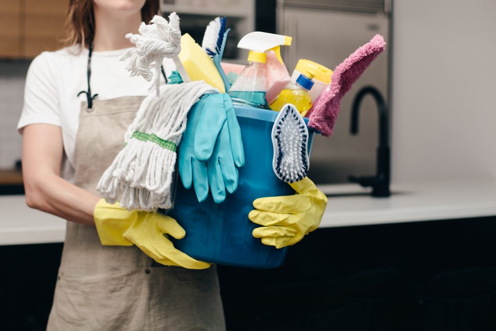 A woman, wearing a tan apron and yellow gloves, holds a blue bucket full of cleaning supplies. 