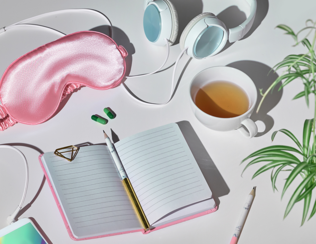 On a white background sits a pink sleep mask in the upper left-hand corner. Next to that are a white pair of corded headphones. Below that is a white teacup with tea in it. To the right of the teacup are green plant leaves. To the left of the plant is a pencil and an open journal with a pencil in it to the left of that. in the bottom left-hand corner is an iPhone that's plugged into a cord.