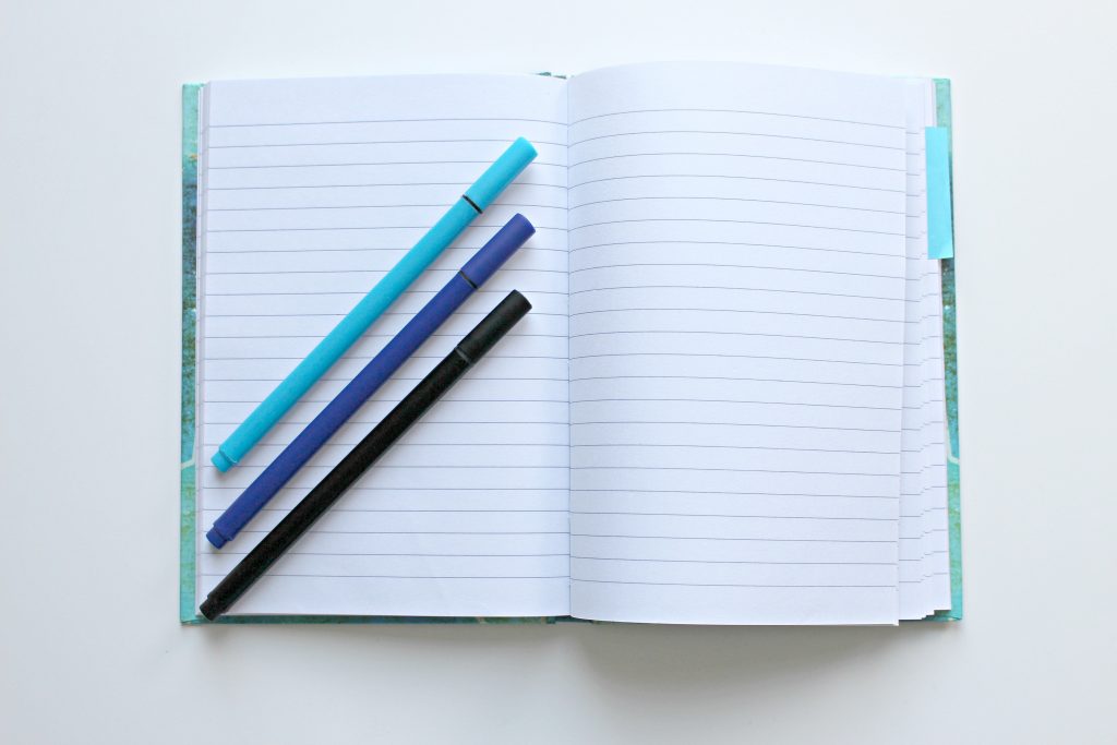 A journal opened to two blank, lined pages. On top of the left page are three pens, light blue, dark blue, and black, sitting on the page at an angle. 