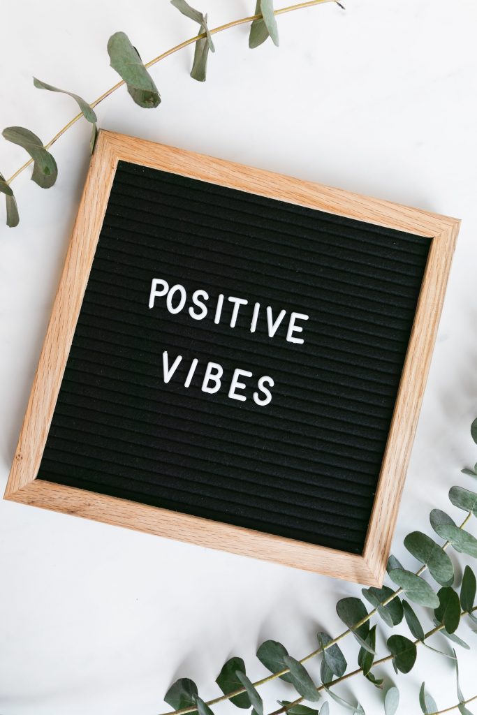 White background with eucalyptus leaves on the top left and bottom right corners. A black letter board with a wooden frame is in the middle with positive vibes in white letters on it. 