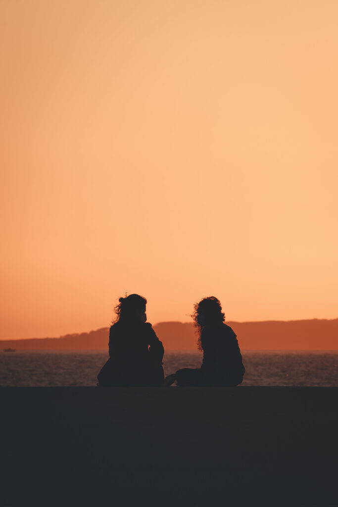 Two women sitting with their backs to the camera. They are in silhouette on a beach, sitting on what looks like black sand, where they will publicly state their goals to each other. There's water past them and the sky is orange from a sunset.  